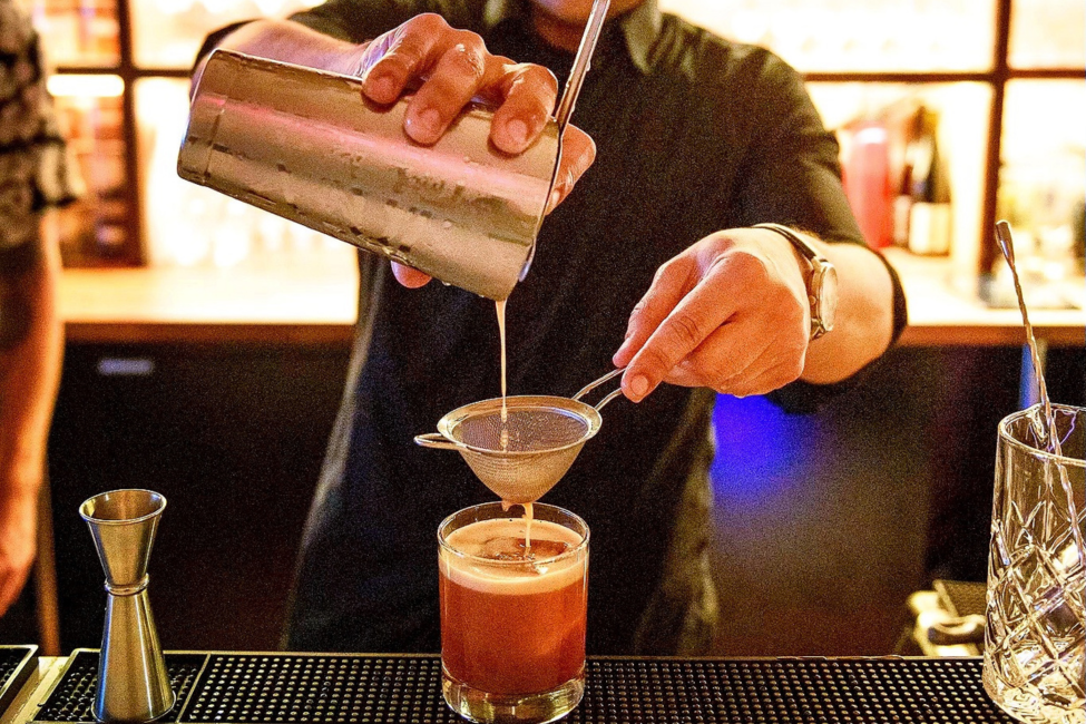 Bartender pouring a cocktail using a strainer and jigger in a bar setting.